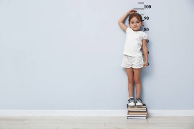 Kitaplarda boyunu beyaz duvarla ölçen sevimli küçük bir kız.