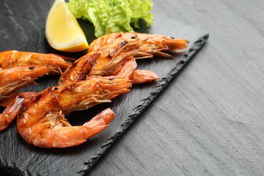 Tasty grilled shrimps with lemon and lettuce on black table, space for text. Delicious seafood dish