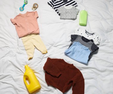 Beyaz çarşafların üzerinde farklı bebek kıyafetleri, oyuncaklar ve deterjanlar var.