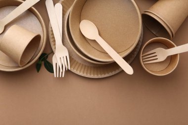 Set of disposable tableware and green leaves on brown background, flat lay. Space for text