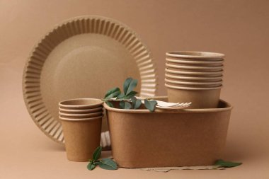 Set of disposable tableware and green leaves on brown background