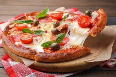 Taking piece of delicious pizza with cheese, salami, tomatoes and olives at wooden table, closeup