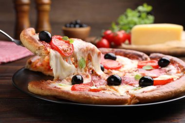 Taking piece of delicious pizza with cheese, salami, tomatoes and olives at wooden table, closeup