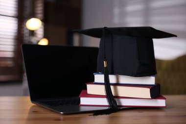 Graduation cap, laptop and stack of books on wooden table, closeup