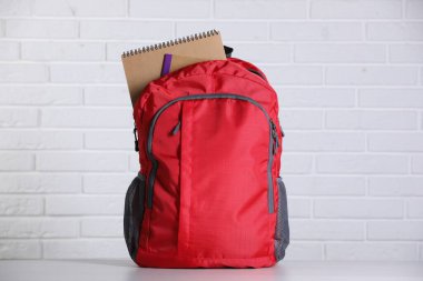 Masanın üzerinde beyaz tuğla duvara karşı defter olan kırmızı sırt çantası.