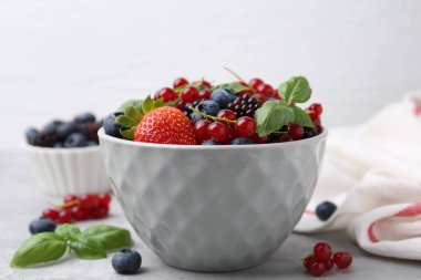 Different ripe berries and basil leaves in bowl on light grey table, closeup