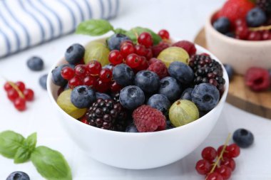 Different ripe juicy berries and basil in bowl on white table, closeup
