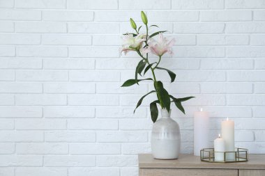Beautiful lily flowers in vase and burning candles on cabinet near white brick wall indoors, space for text