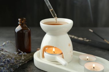 Dripping essential oil from pipette into aroma lamp with burning candle at grey textured table, closeup