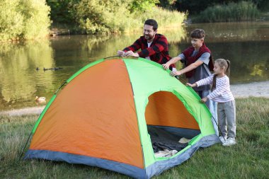 Mutlu baba ve çocukları nehir kenarında kamp çadırı kuruyorlar.