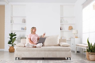 Dizüstü bilgisayarı olan bir kadın evdeki koltukta klimayı kontrol etmek için uzaktan kumanda kullanıyor.