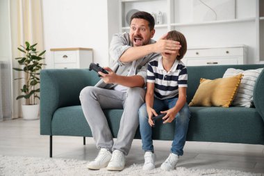 Evde kanepede video oyunu oynarken kontrolörlerle eğlenen baba ve oğul.