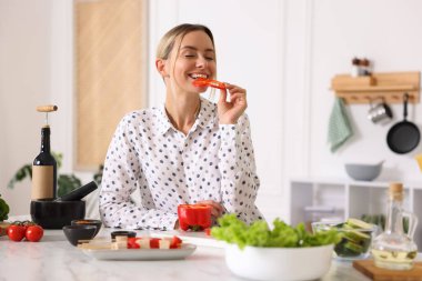 Mutfakta yemek pişirirken biber yiyen gülümseyen kadın.