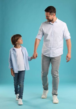 Baba oğul açık mavi arka planda yürürken el ele tutuşuyorlar.