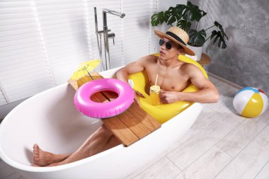 Güneş gözlüklü ve hasır şapkalı genç adam evdeki küvette şişme yüzüklü mason kavanozundan meyve suyu içiyor. Denizde tatilin hayalini kuruyorum.