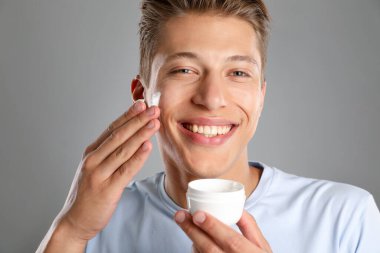 Yakışıklı genç adam gri arka planda yüzüne krem sürüyor.