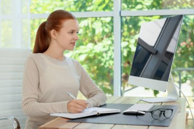 Genç kız bilgisayarını kullanıyor ve içerideki ahşap masada deftere yazı yazıyor. Ev işyeri