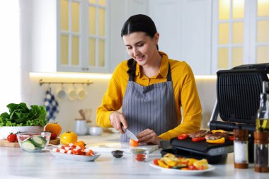 Kadın mutfakta elektrikli ızgarada et ve sebze pişirirken biber kesiyor.