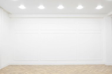 Beyaz duvarlı boş oda, ahşap zemin ve tavanda benekli lambalar.