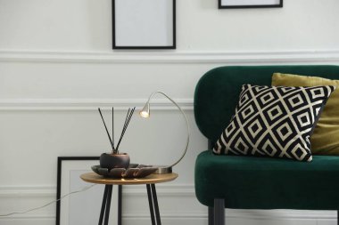 Stylish lamp and reed diffuser on side table near armchair with pillows indoors