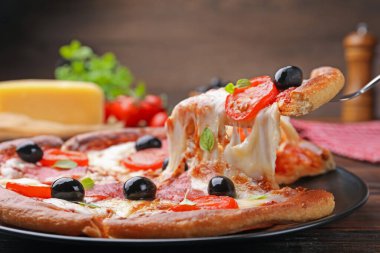 Taking piece of delicious pizza with cheese, salami, tomatoes and olives at wooden table, closeup