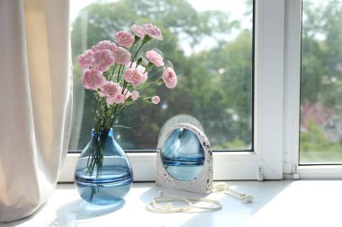 Beautiful carnation flowers in vase, mirror, jewellery and face roller near window indoors