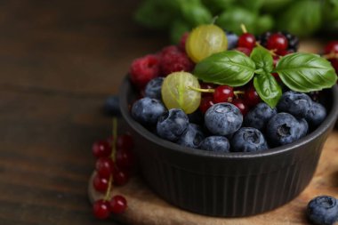 Different ripe juicy berries and basil in bowl on wooden table, closeup. Space for text