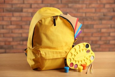 Sınıftaki masanın üstünde not defterleri ve sanat malzemeleri olan sarı sırt çantası, yakın plan.