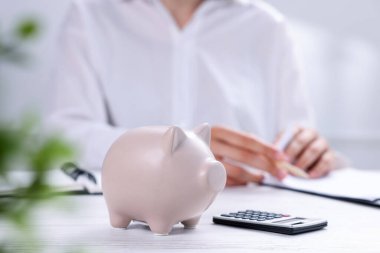 Beyaz ahşap masa üzerinde hesap makinesi olan domuz kumbarası ve kapalı alanda çalışan kadın, seçici odak