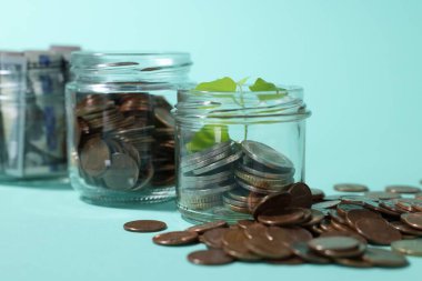 Bozuk paralı cam kavanozlar, filiz ve açık mavi arka planda dolar, yakın plan.