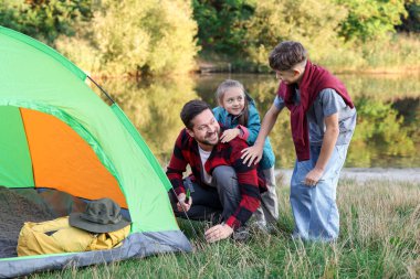 Mutlu baba ve çocukları nehir kenarında kamp çadırı kuruyorlar.