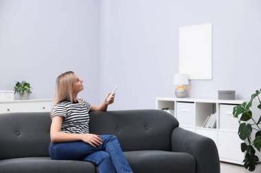 Evdeki kanepede uzaktan kumandayla klima kullanan bir kadın. Metin için boşluk