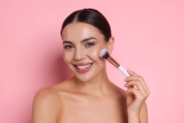 Güzel bir kadın pembe arka planda fırçayla makyaj yapıyor.