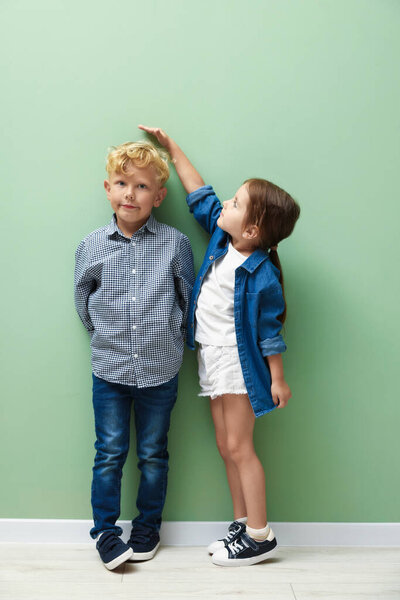Cute little girl and boy measuring their heights near green wall