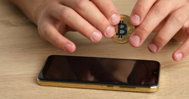 Kripto para birimi. Elinde altın bitcoin ve akıllı telefon olan adam ahşap masada, yakın plan.