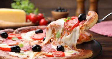 Taking piece of delicious pizza with cheese, salami, tomatoes and olives at wooden table, closeup