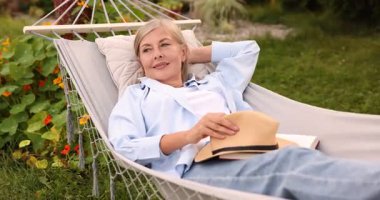 Hasır şapkalı ve kitaplı yaşlı kadın dışarıda hamakta dinleniyor.