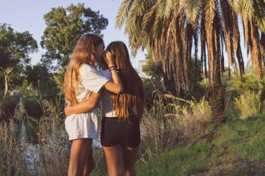 Two teen girls embracing in a park, one whispering a secret to the other, viewed from behind