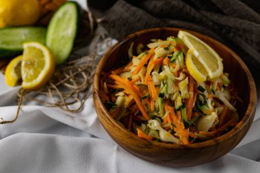 Derin bir tabakta taze sebzelerden oluşan sağlıklı bir salata. Beyaz lahana, havuç ve limon dilimleri ve otlarla süslenmiş mis kokulu salatalıkla dolu bir tabak. Sağlıklı beslenmenin ilginç bir sunumu