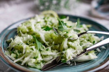 Taze sebzeli çıtır salata. Limonla süslenmiş salatalık ve roka yapraklı lahana ve çatal bıçaklı, hafif desenli taş bir masada bir buket beyaz çiçek.