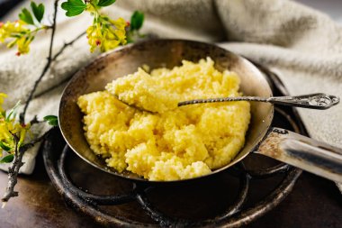 Eski bir tavada şeker ve tereyağlı mısır lapası ve eski bir metal şarap standı. Yanında sarı çiçekli, çiçek açan siyah bir frenk üzümü var.