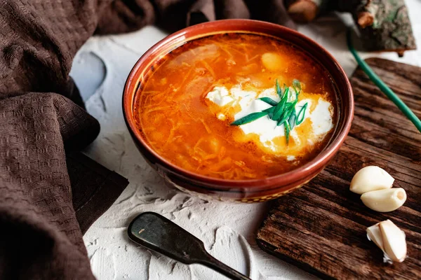A portion of red borscht in a brown plate, next to an old wooden Board garlic and quail egg, green leeks and driftwood. White sour cream in the soup, next to an old Polish spoon — Stock Image Portion Red Borscht Brown Plate Next Old Wooden Board Garlic Stock Photo