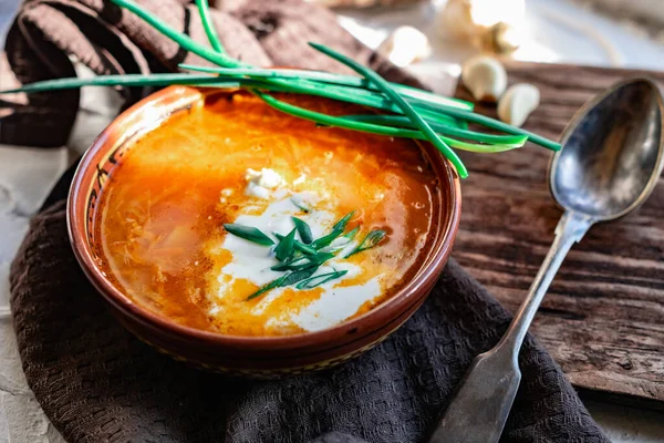 A portion of red borscht in a brown plate, next to an old wooden Board garlic and quail egg, green leeks and driftwood. White sour cream in the soup, next to an old Polish spoon — Stock Image Portion Red Borscht Brown Plate Next Old Wooden Board Garlic Stock Image