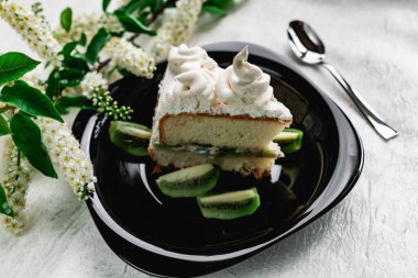 Parlak siyah bir tabakta taze kivi dilimli kremalı bir dilim pasta. Pasta, mis kokulu kiraz çiçekleriyle süslenmiş.