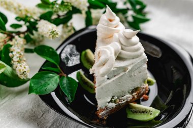 Parlak siyah bir tabakta taze kivi dilimli kremalı bir dilim pasta. Pasta, mis kokulu kiraz çiçekleriyle süslenmiş.