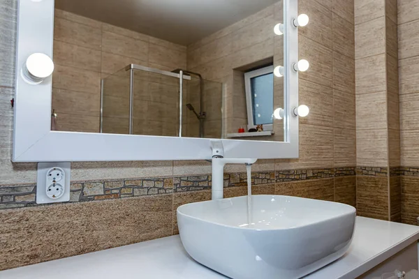 The interior of the bathroom with a big illuminated mirror, walk-in shower and elegant sink