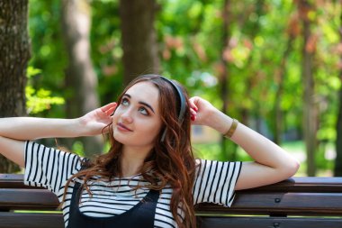 Genç, güzel, kızıl saçlı bir kız bir bankta otururken kulaklıkla müzik dinliyor.