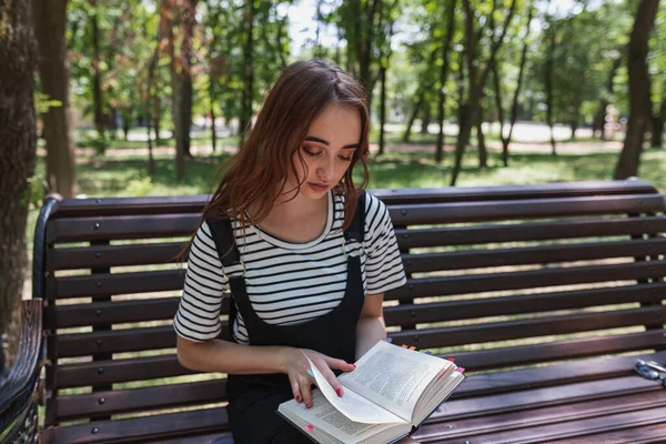 Çekici kızıl saçlı genç kız parkta kitap okuyor ve sınava hazırlanıyor.