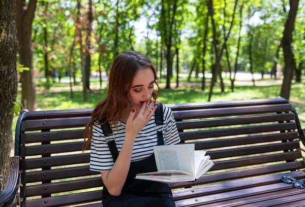 公園の魅力的な赤髪の10代の少女はベンチで本を読んでいます 彼は指をクリックして本のページをめくる — ストック写真