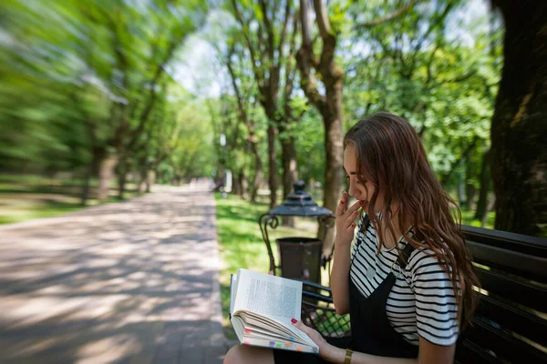 公園の魅力的な赤髪の10代の少女はベンチで本を読んでいます 彼は指をクリックして本のページをめくる — ストック写真